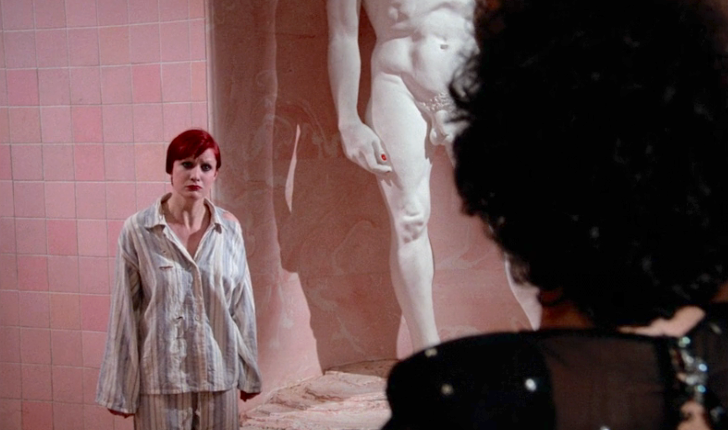 A person in pajamas stands facing a large classical-style statue. Another person's silhouette is in the foreground