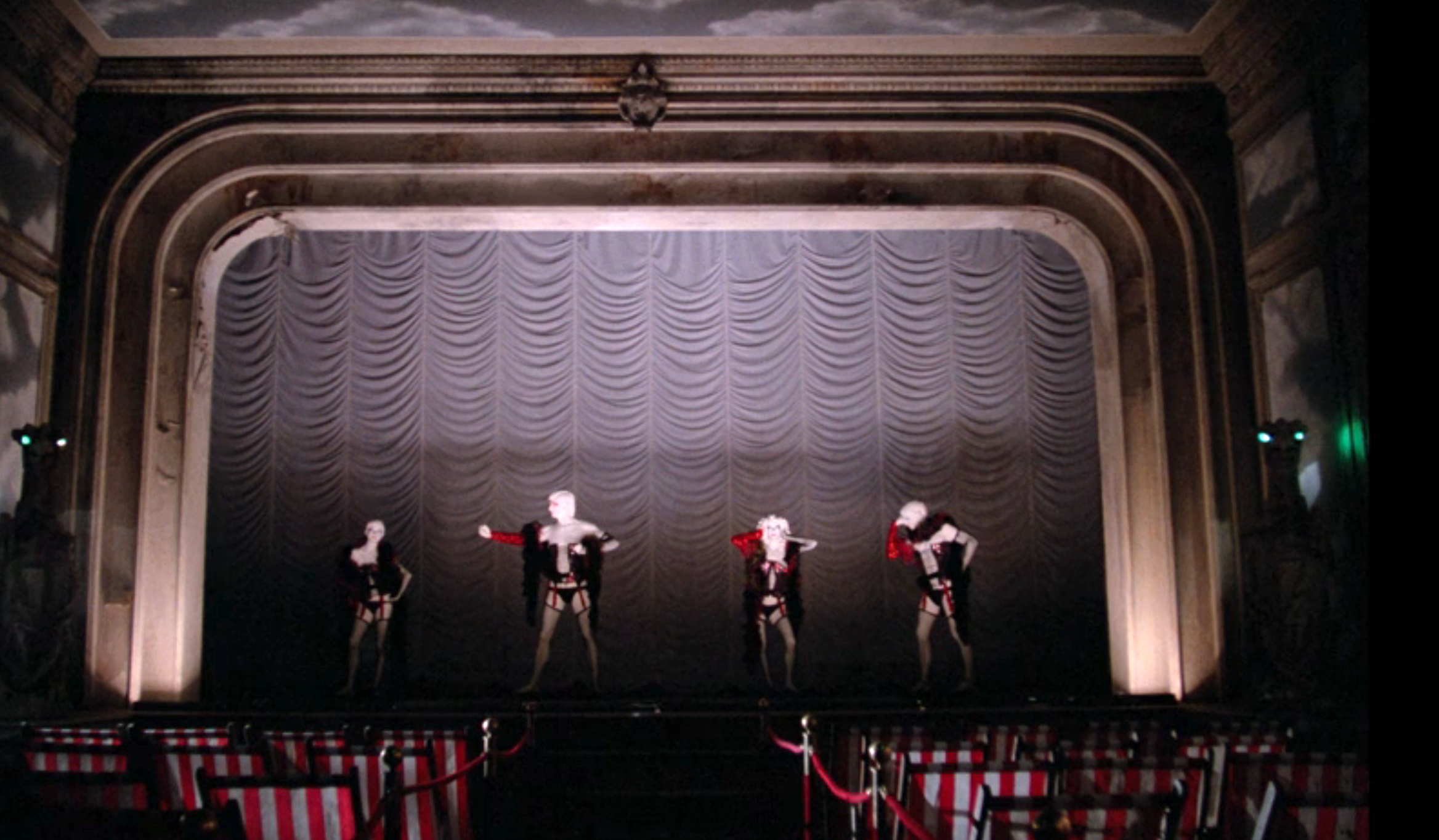 Three performers in theatrical costumes pose on an ornate stage with a curtain backdrop, suggesting a cabaret or theatrical show setting