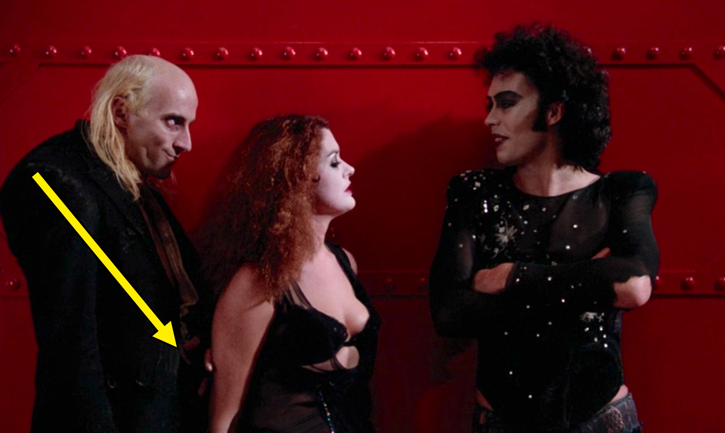 Three people in theatrical gothic costumes and makeup stand against a red wall. The central figure has voluminous curly hair and a low-cut outfit