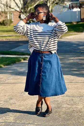striped sweater, buttoned denim skirt, loafers, and ample earrings, lasting with hands adjusting glasses