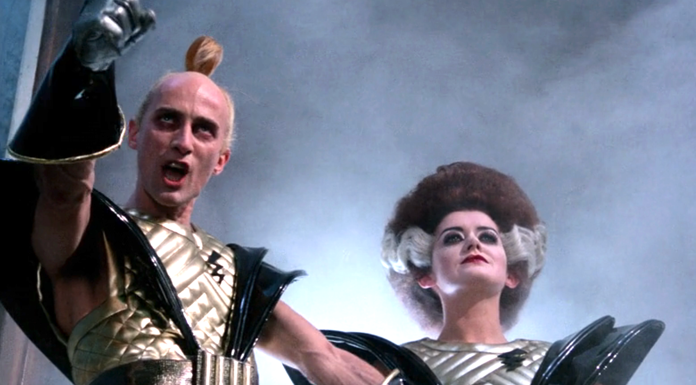 Two performers in futuristic costumes, one with a mohawk and metallic armor, the other with striking makeup and a voluminous hairstyle, pose dramatically