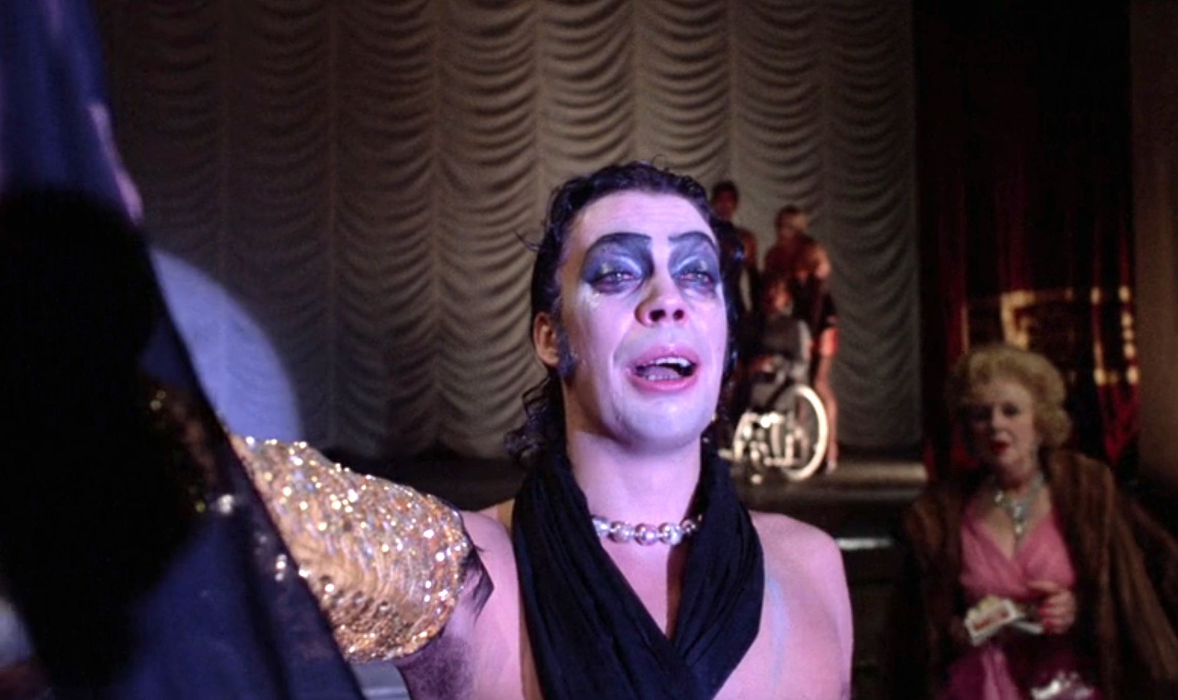 Person in theatrical makeup and sequin outfit performing on stage with others in the background, one in a wheelchair