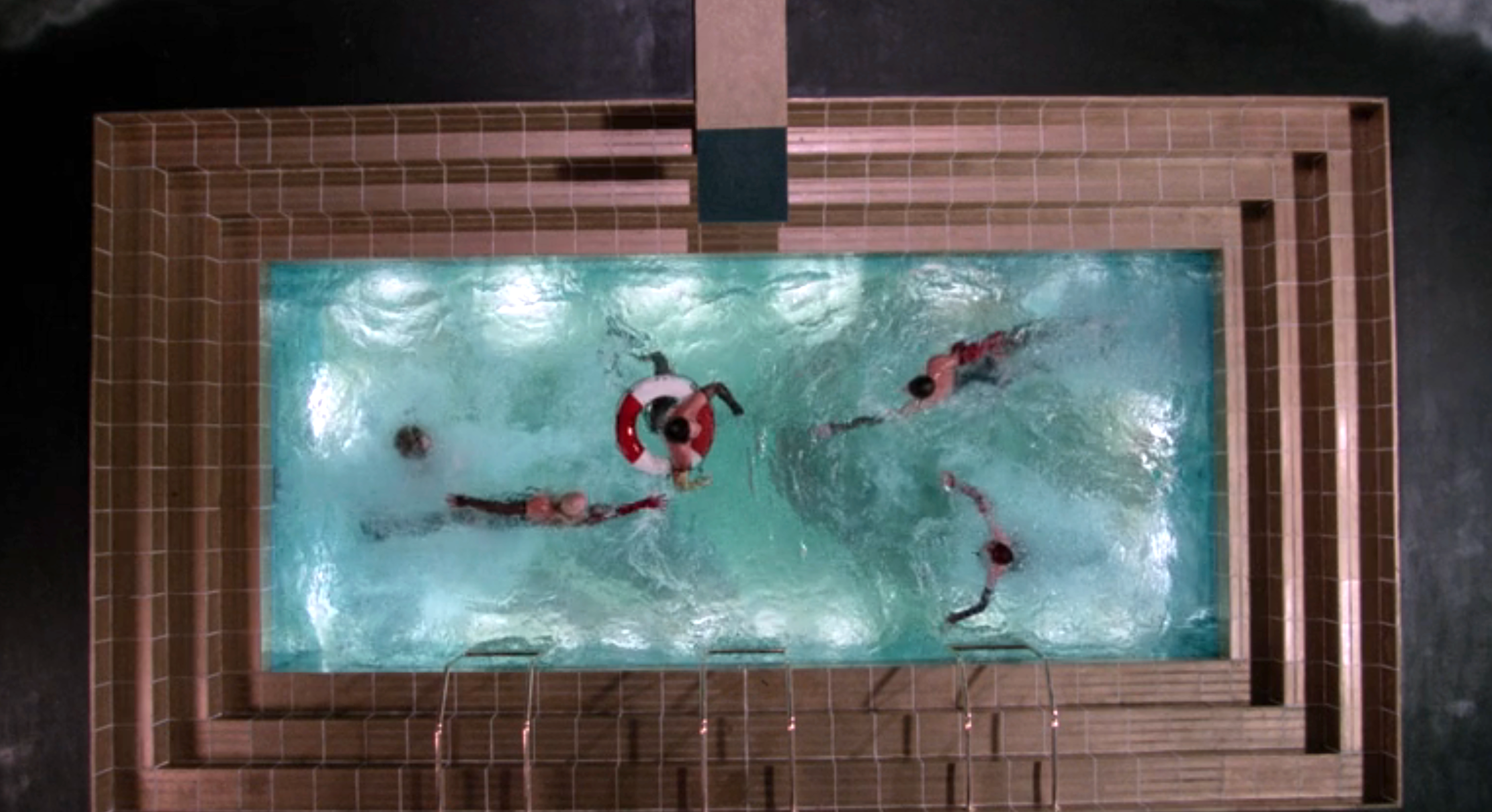 Top-down view of people swimming in a glass-bottom pool, built over a high structure. One person floats with a lifebuoy