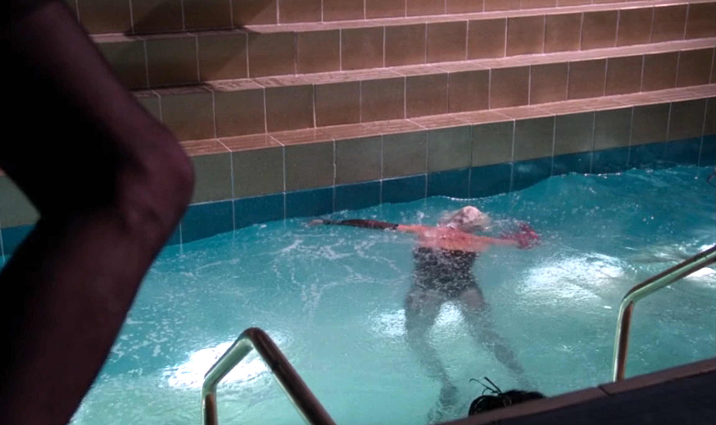 Person swimming alone in an indoor pool, performing a backstroke under low lighting. Steps and railing visible at pool edge