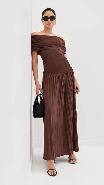 Person successful a brownish off-shoulder, pleated maxi dress, achromatic handbag, and sunglasses, leaning against a partition successful a fashion-forward pose