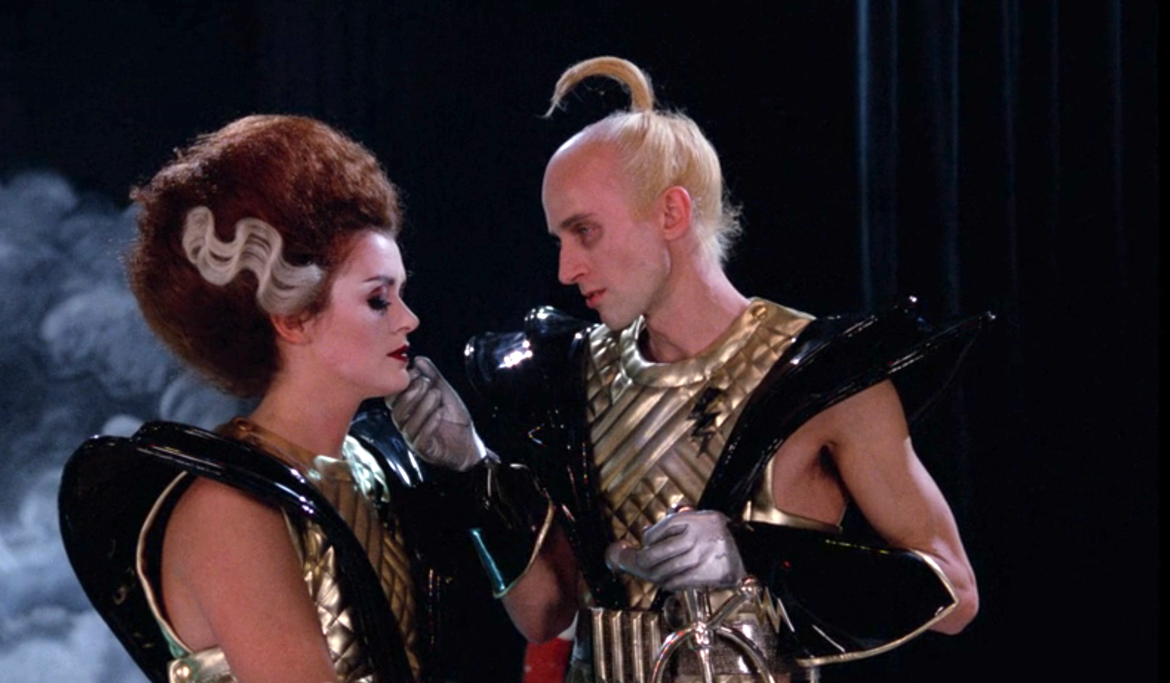 Two people in futuristic, avant-garde outfits with metallic accents, standing closely and interacting on stage
