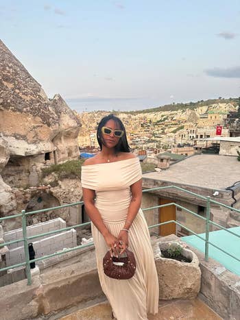 Person wearing an off-shoulder gown and sunglasses stands against a scenic rocky landscape, holding a tiny bag