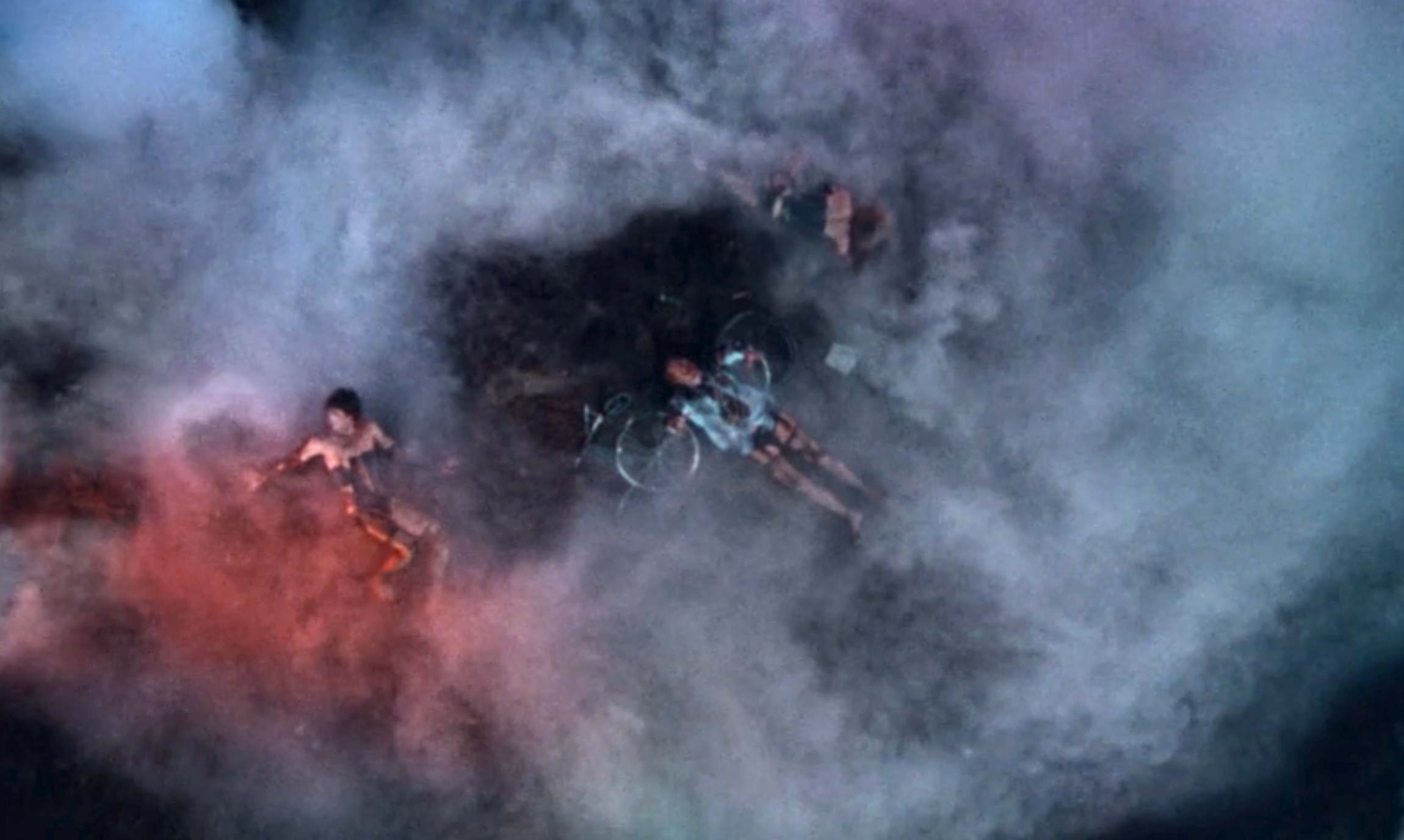 Three people appear to be lying on the ground surrounded by smoke, with a bicycle nearby, suggesting a mysterious or dramatic scene