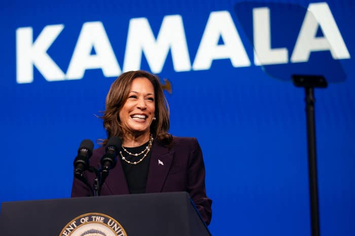 A person stands smiling at a podium with "KAMALA" in large text behind them. They are wearing a dark suit and a layered necklace