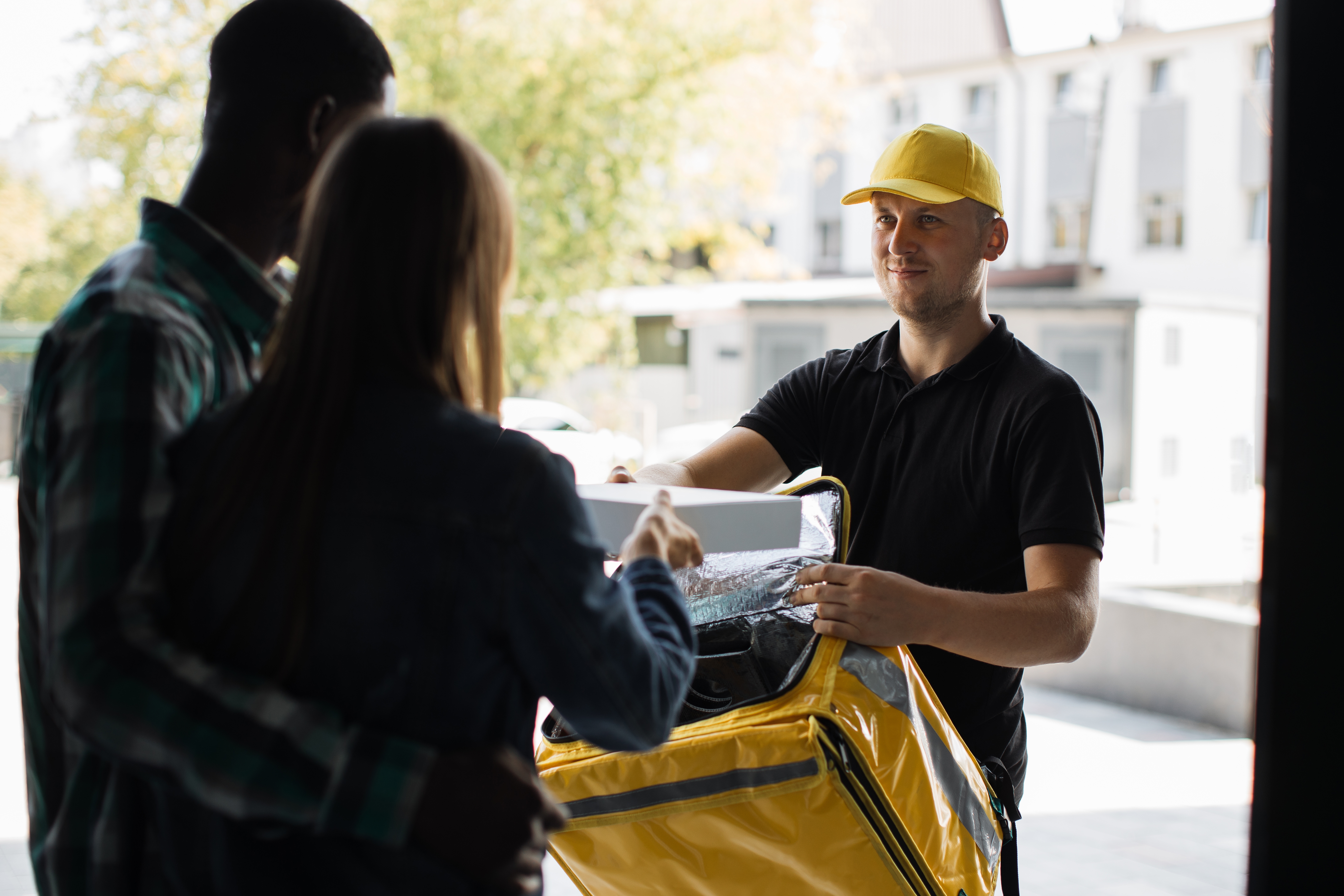 Delivery worker hands package to a couple at a door. Worker wears a cap and holds an insulated bag