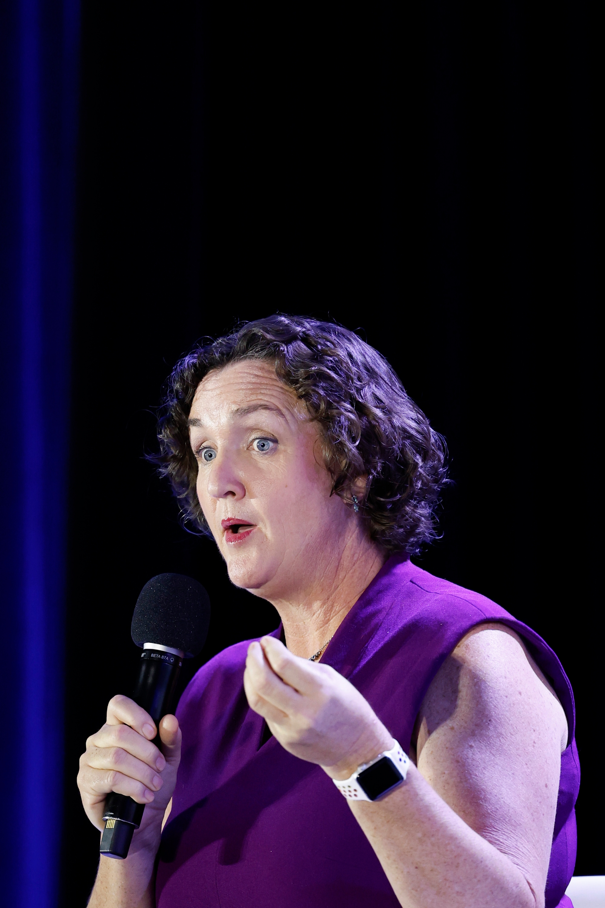 Person speaking onstage, holding a microphone. They are wearing a sleeveless dress and a smartwatch, gesturing with one hand