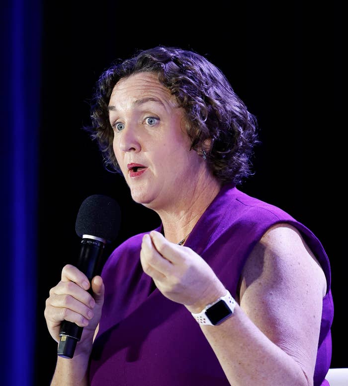 Person speaking onstage, holding a microphone. They are wearing a sleeveless dress and a smartwatch, gesturing with one hand