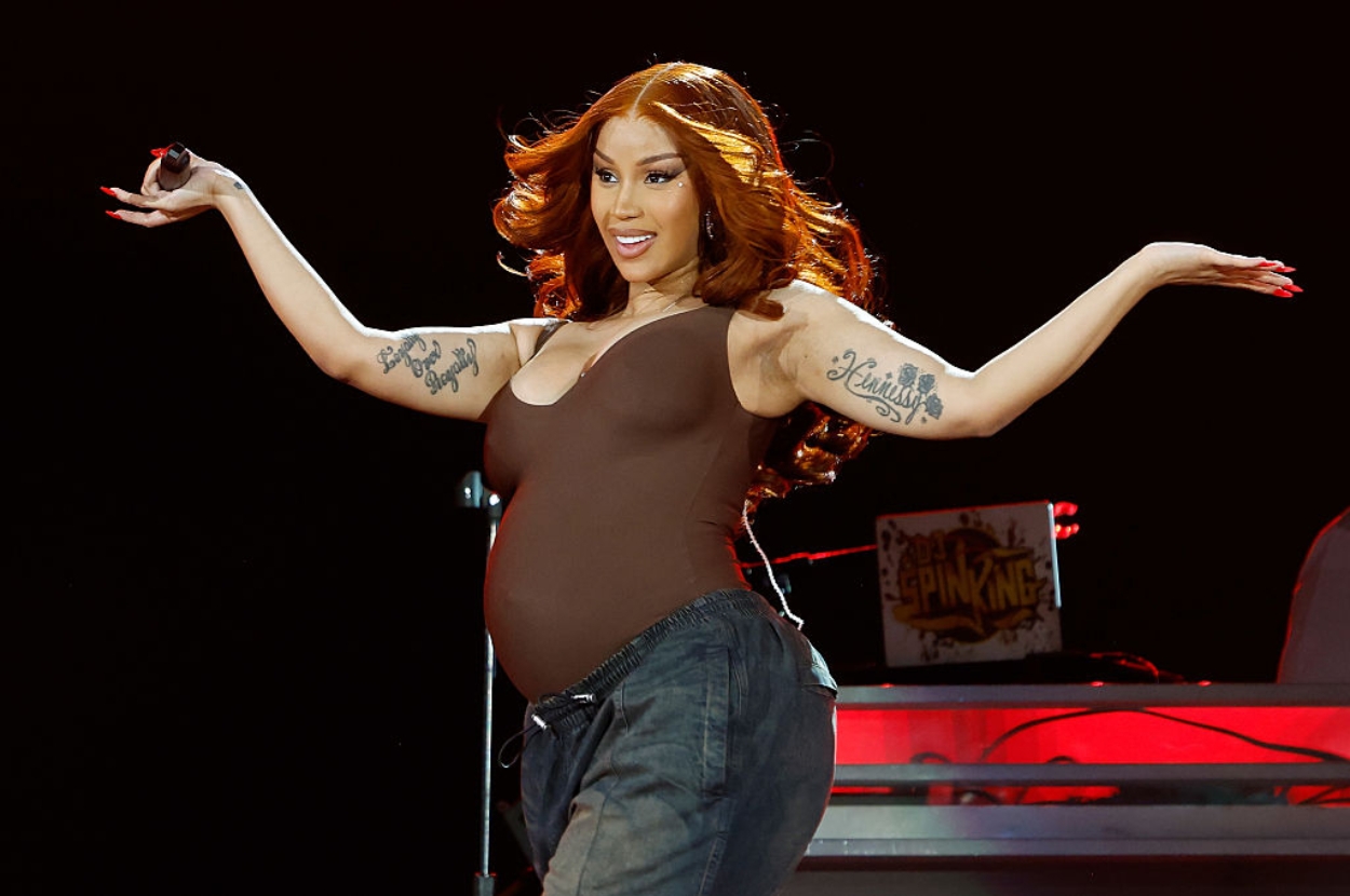 Pregnant performer on stage, smiling with arms out, wearing a fitted top and relaxed pants, showcasing tattoos