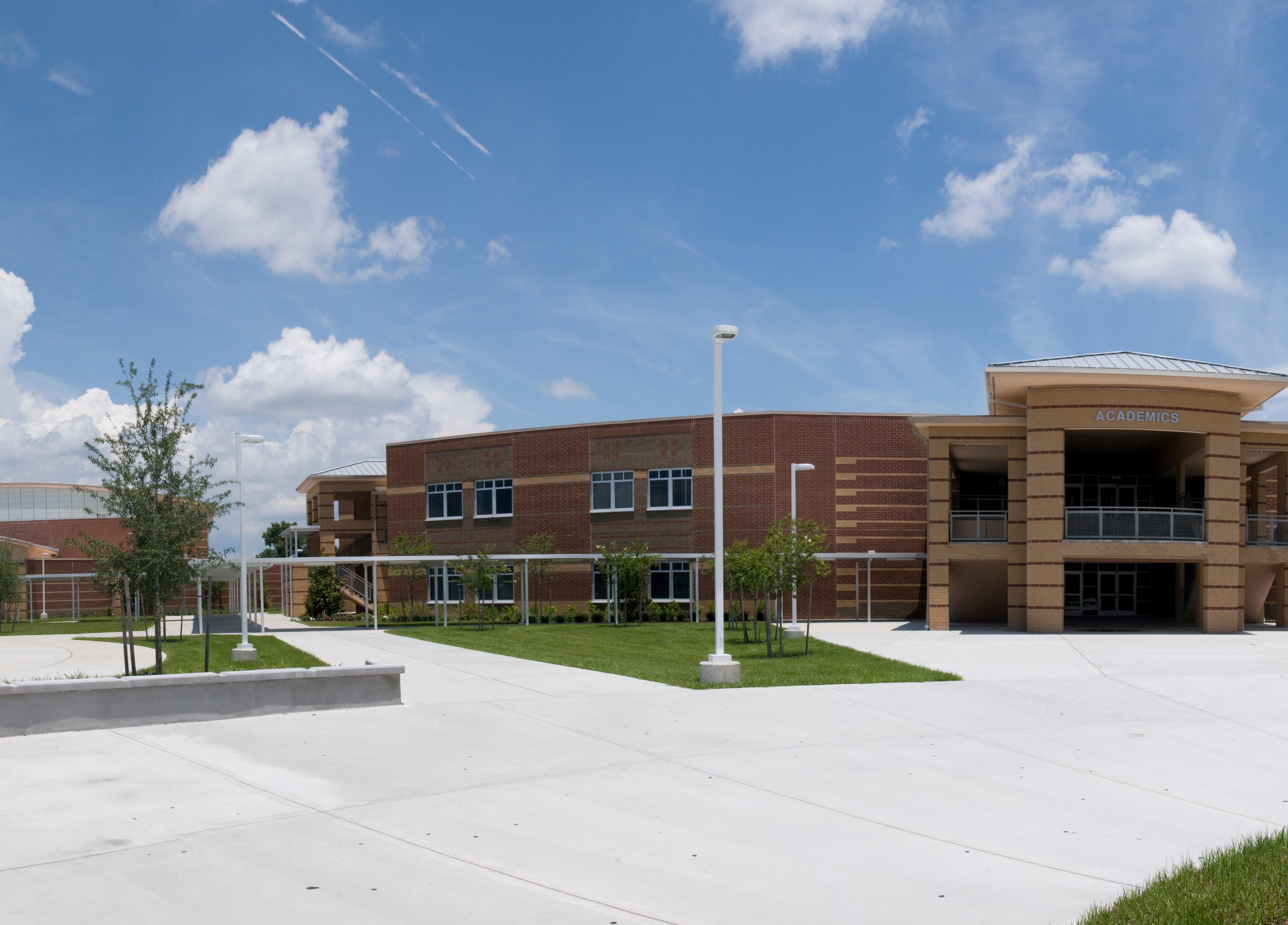 Moderne skolebygg med store vinduer, omgitt av en romslig asfaltert gårdsplass og få trær under delvis overskyet himmel