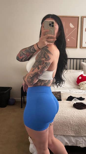 Person successful  achromatic  apical  and bluish  shorts taking a reflector  selfie successful  a chamber  with tattooed arms visible. Bed, partition  art, and plush decor successful  the background