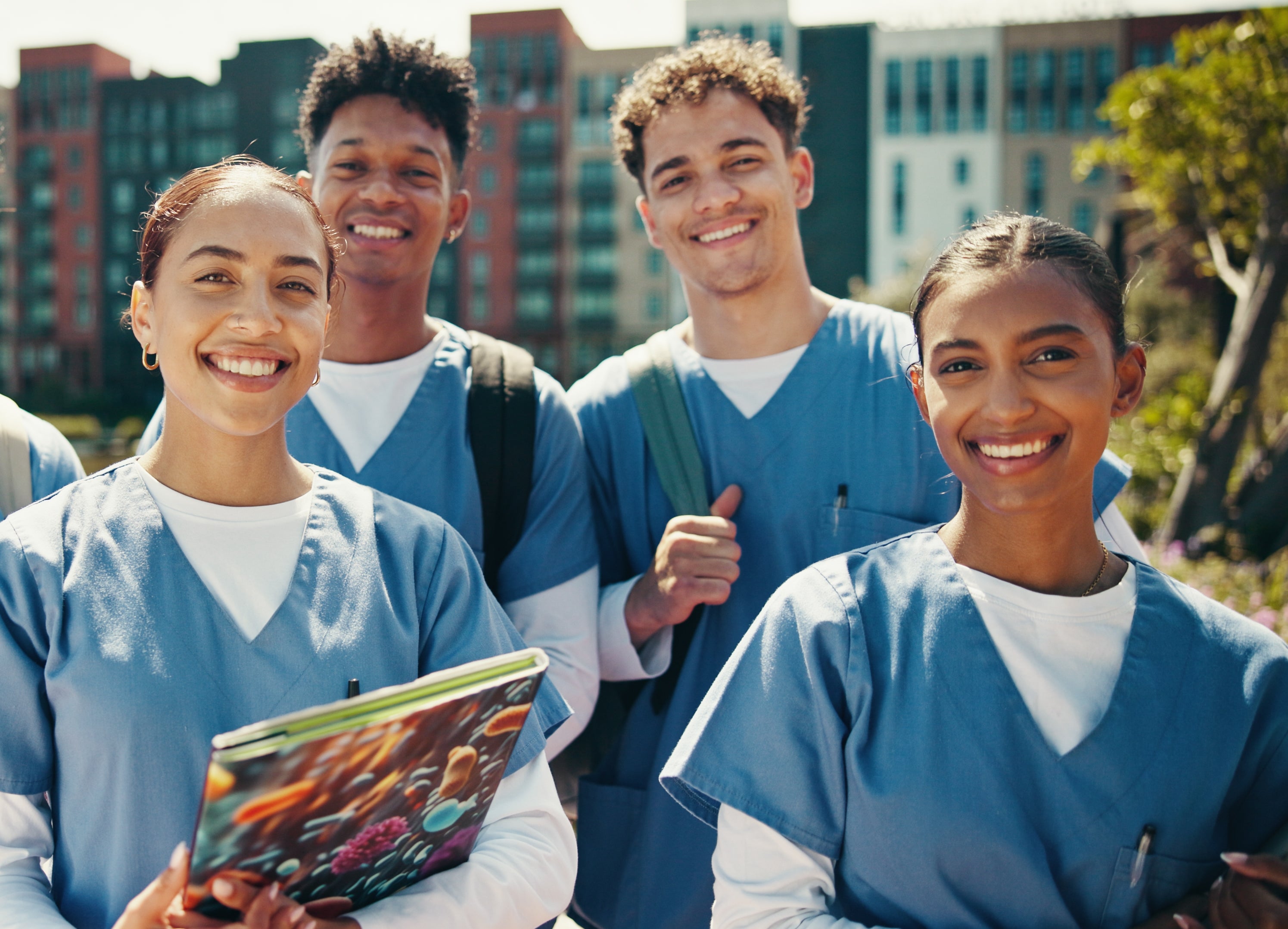 Fem smilende medisinstudenter i skrubb, holder nettbrett og notatbøker, står utendørs i urbane omgivelser