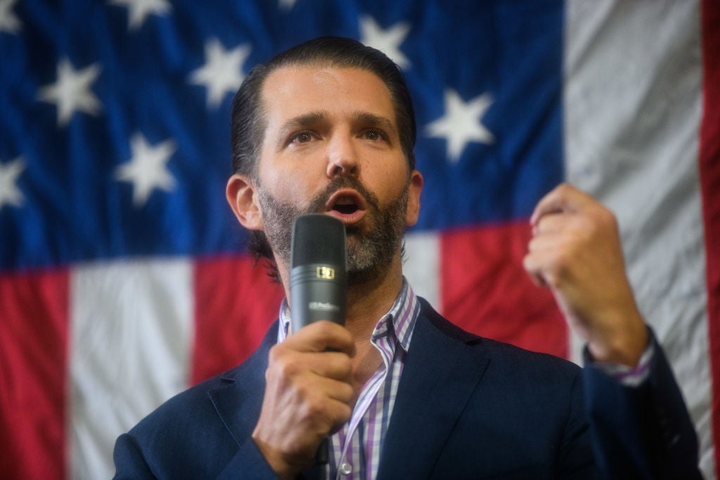 Person speaking passionately into a microphone, standing in front of a large American flag backdrop