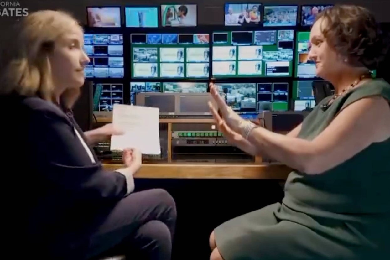 Two women sit across from each other in a TV control room, one holding papers, the other gesturing with hands raised. Monitors display varied content