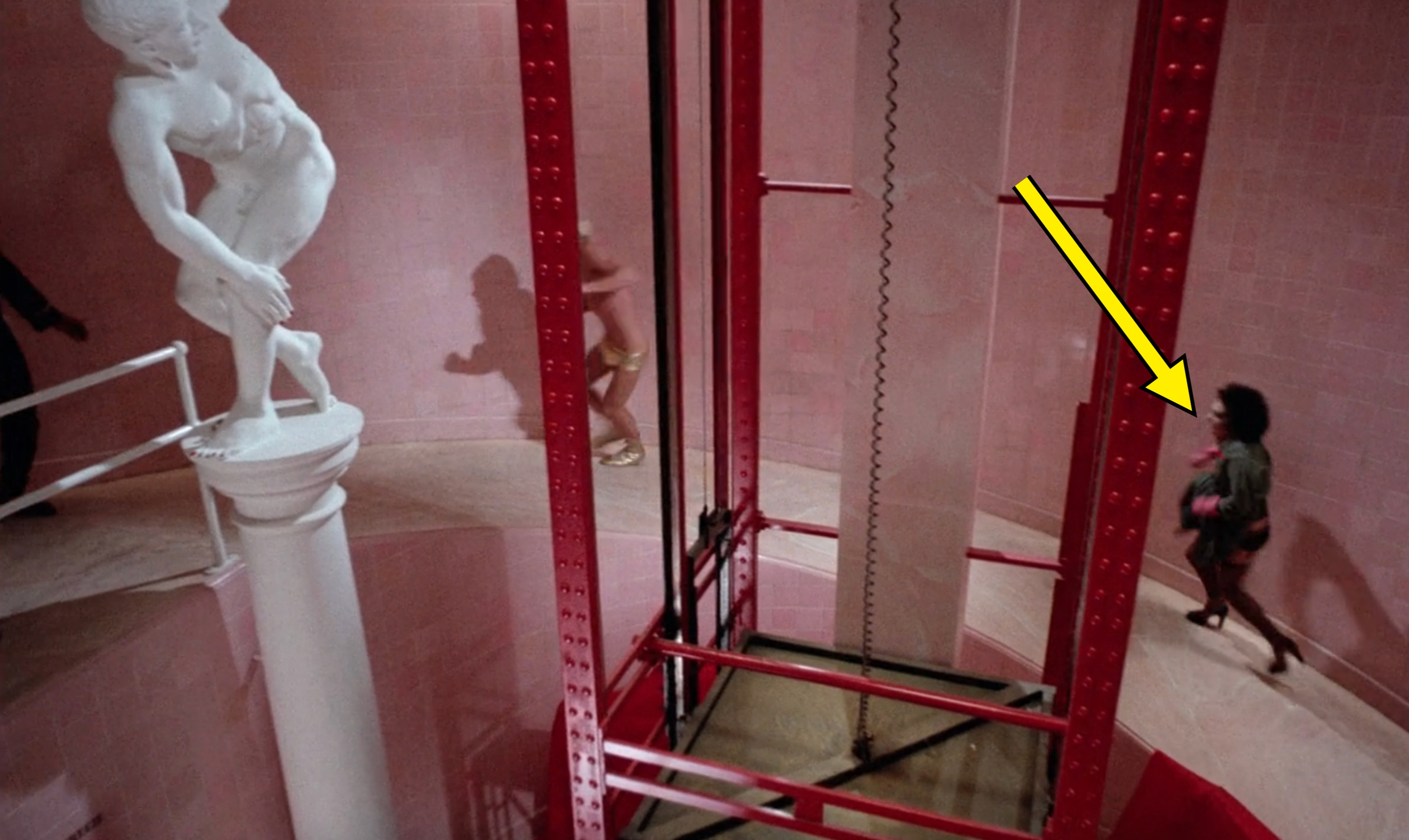 Sculpture of a crouching figure on a pedestal, with people walking around a circular red structure in a modern architectural space