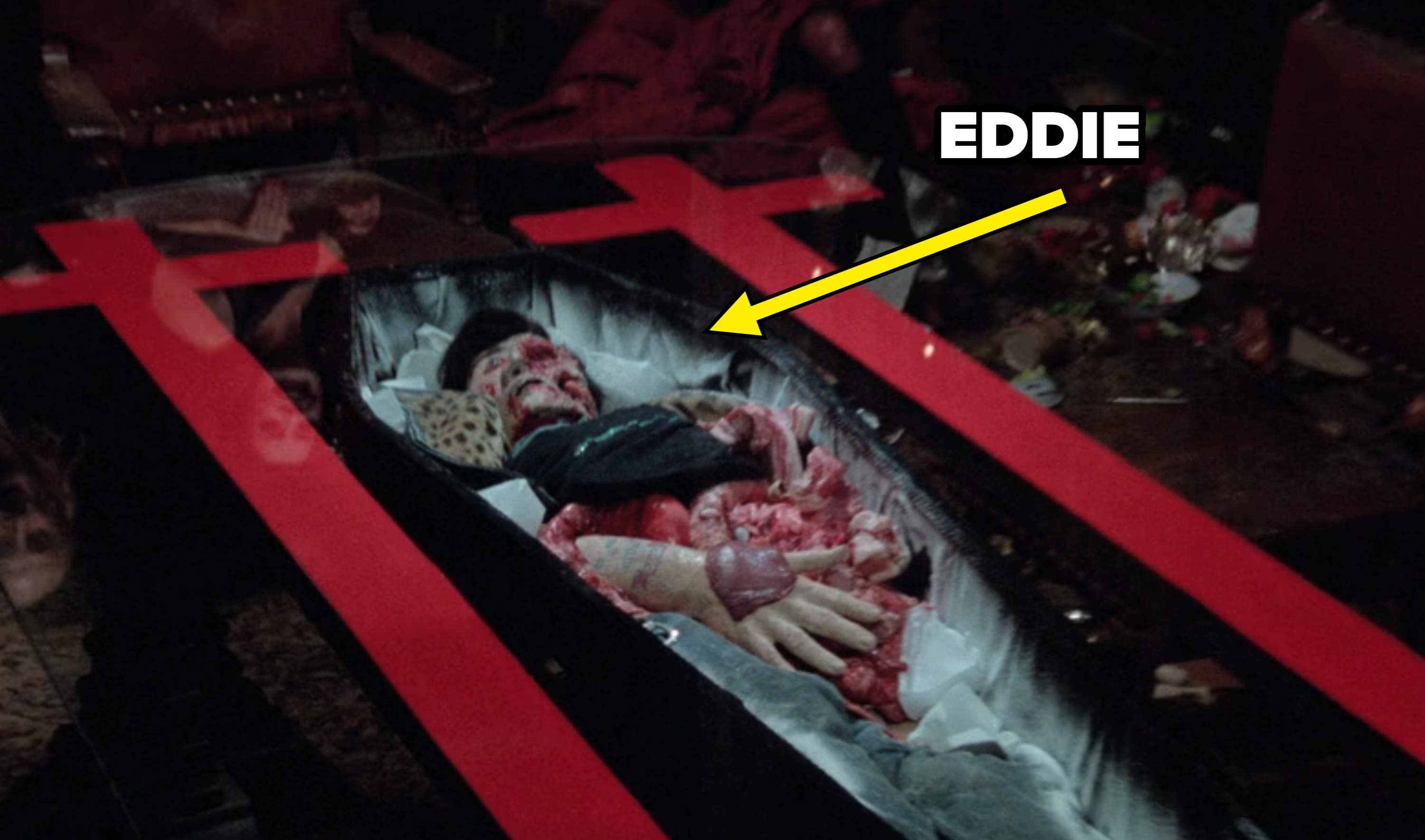 A scene from a movie depicting a character with visible fake gore and prosthetics, lying in a casket with red crosses on the glass lid