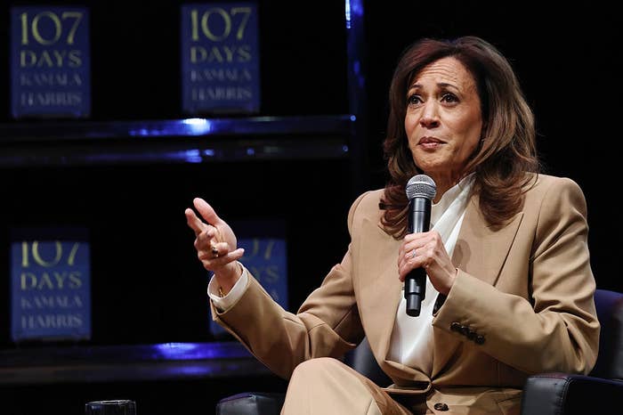 Person speaking on stage with microphone, wearing a brown suit seated in front of shelves filled with books titled "107 Days."
