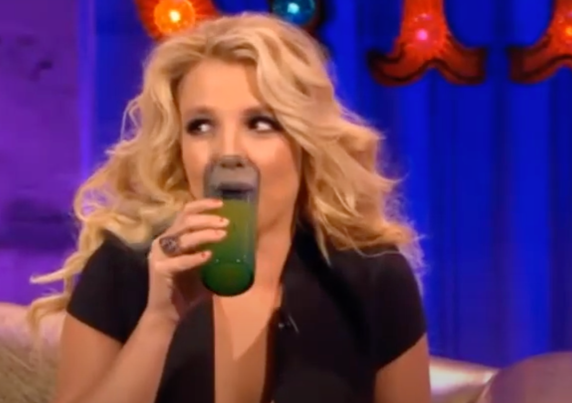 Personen smiler mens han holder en grønn drink. De sitter på en sofa i det som ser ut som en livlig setting