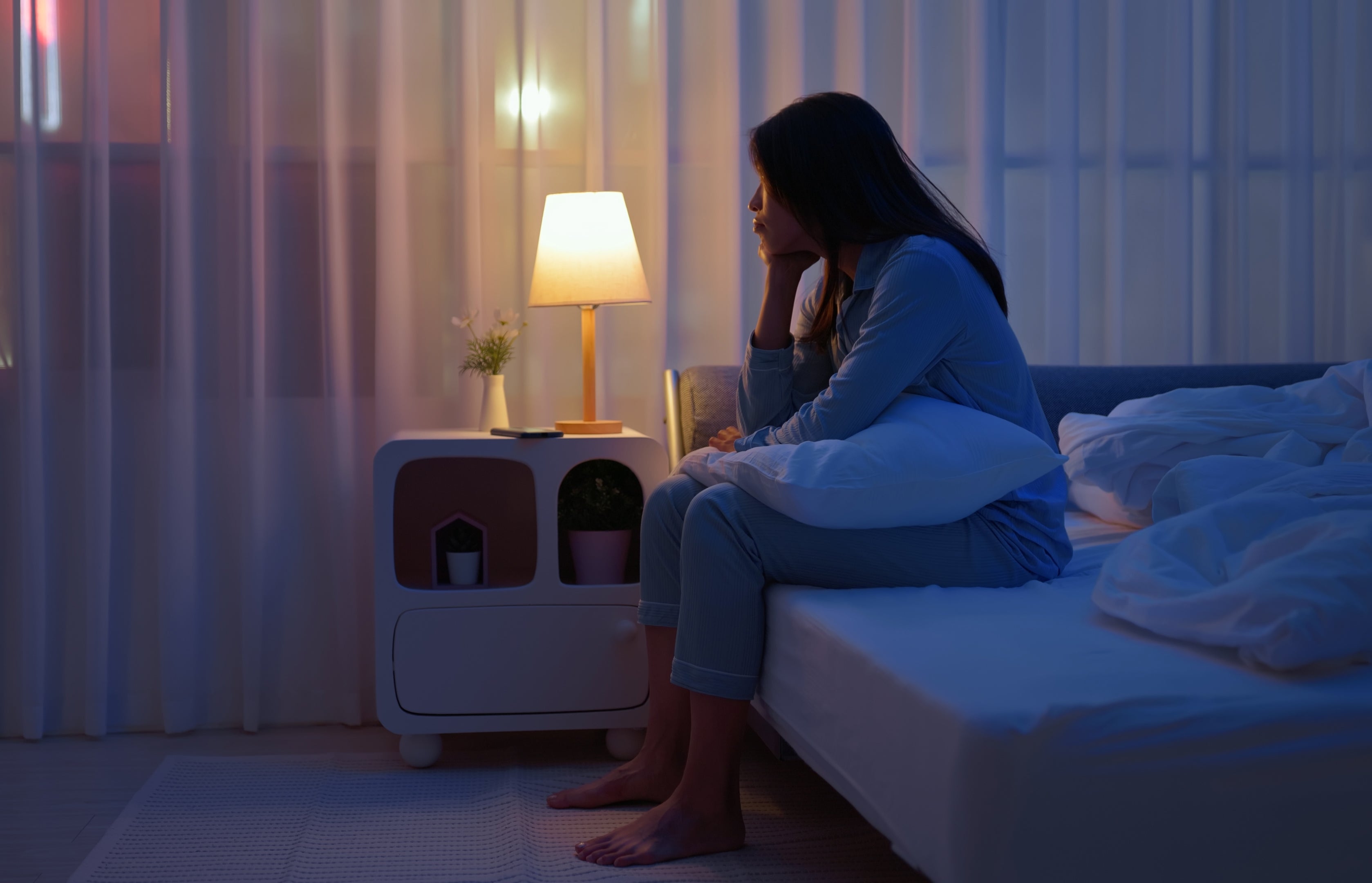 En person i pyjamas sitter pensivt på en seng om natten og holder en pute, med en tent lampe på nattbordet