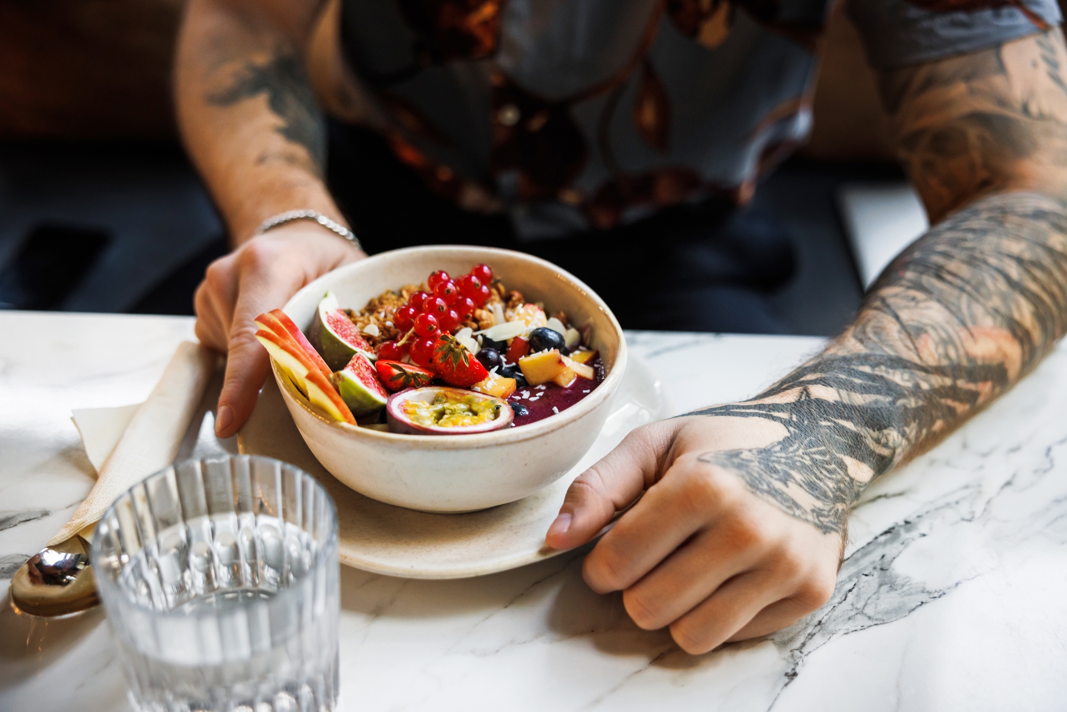 Person med tatoveringer som holder bolle med smoothie toppet med frukt og frø, sitter ved et marmorbord med et glass vann