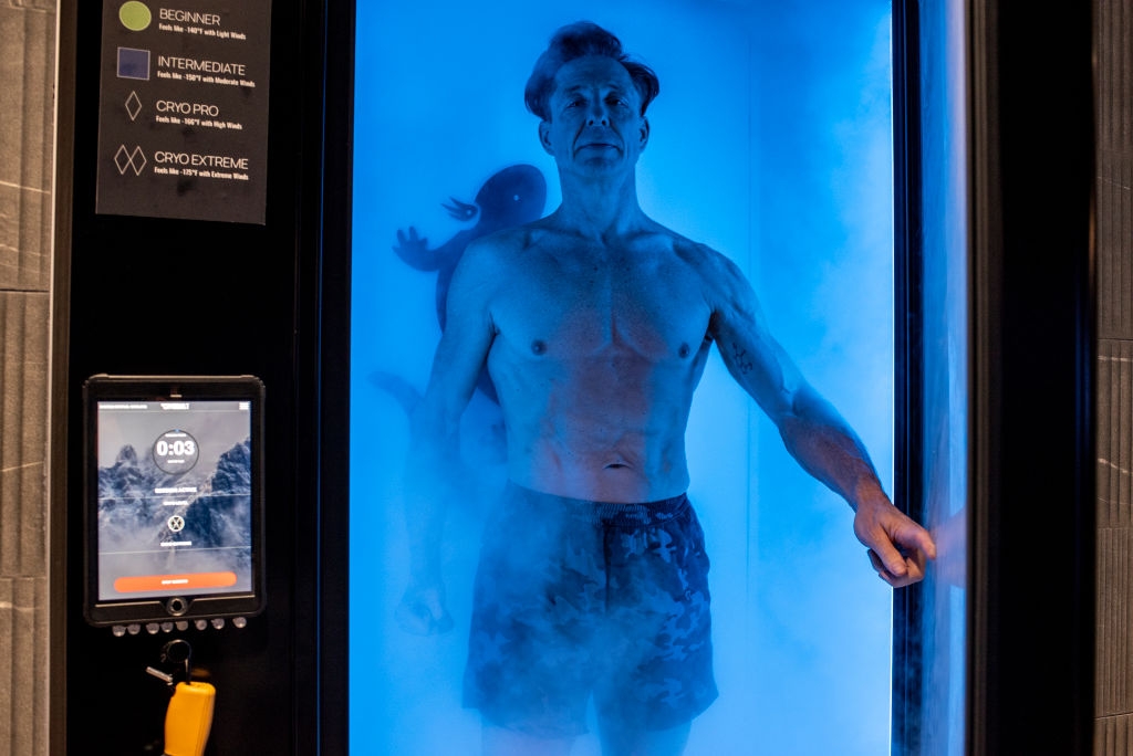 En mann står i et kryoterapikammer, delvis skjult av tåke, iført shorts. En digital skjerm viser alternativer utenfor kammeret