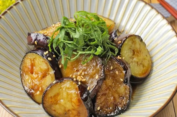 白い器に盛られたナスの料理。上に細く刻んだ薬味がのっており、ごまが散らされています。テーブルに箸が置かれています。