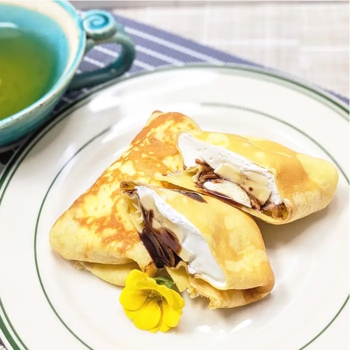 お皿に盛られたクレープの中にバナナとチョコレートが見える。横に小さな黄色い花が飾られている。