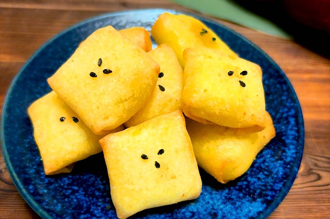お皿に盛られた焼き菓子のイメージ。四角い形で、ごまが上にトッピングされています。背景にさつまいもが見えます。