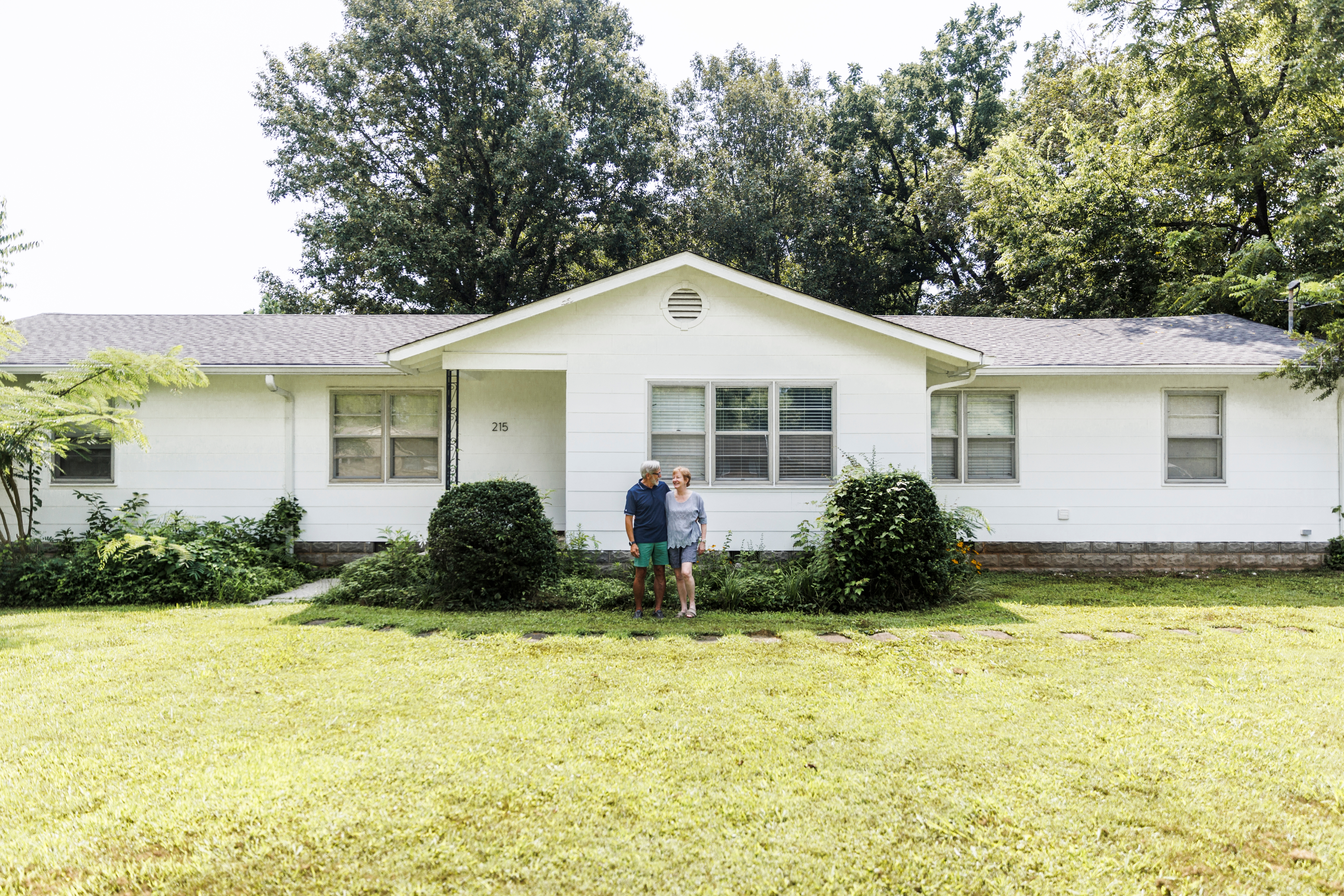 Et par står foran et forstadshus, omgitt av grøntområder og trær, på en solskinnsdag