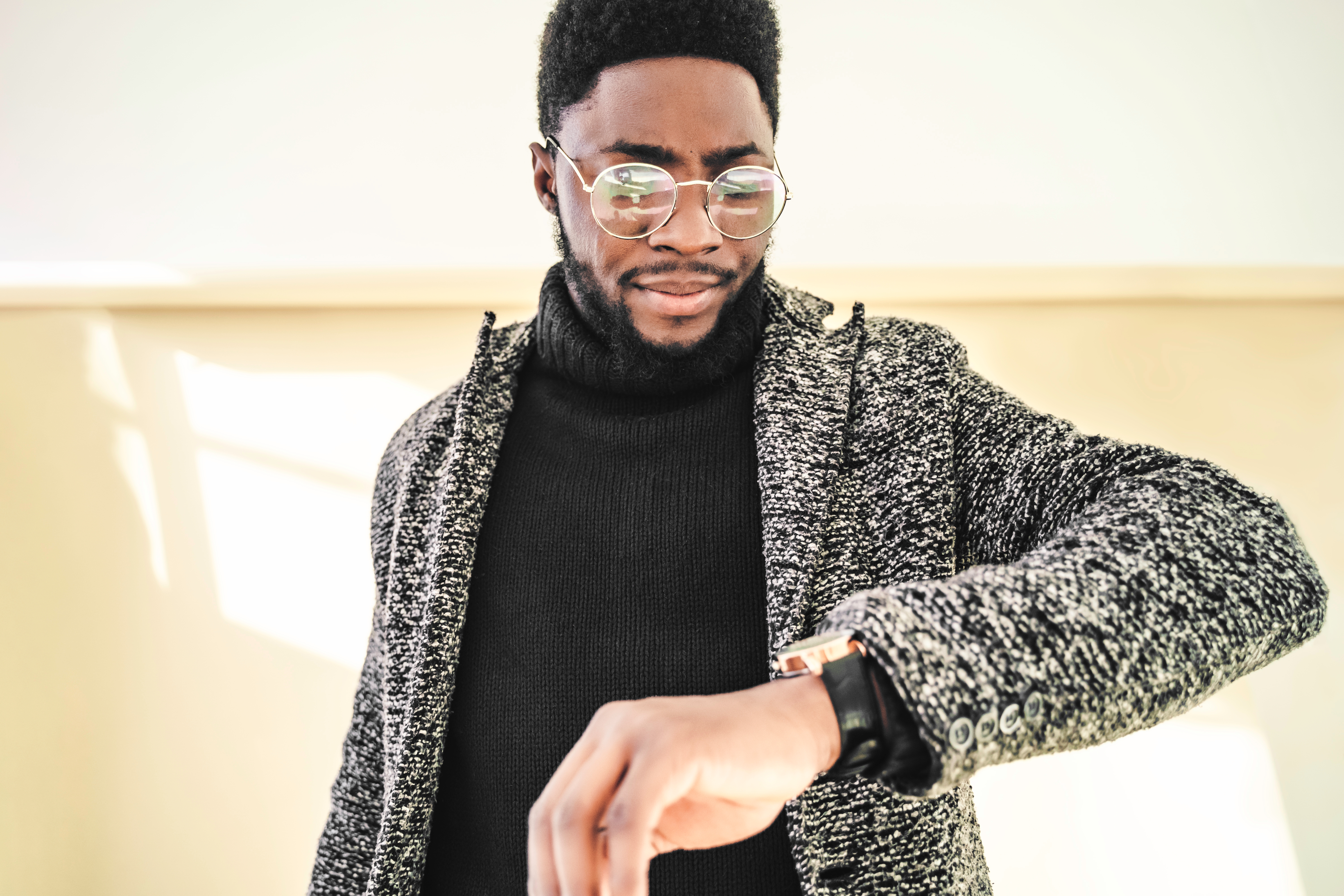 Mann i en strukturert jakke og briller som sjekker en klokke på håndleddet, stående innendørs med nøytral bakgrunn