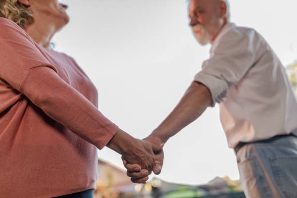 Two people hold hands tenderly, suggesting a romantic connection. They are outdoors, one wearing a casual sweater, the other in a rolled-up shirt