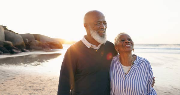 Older couple smiling and embracing on a beach at sunset, conveying love and companionship