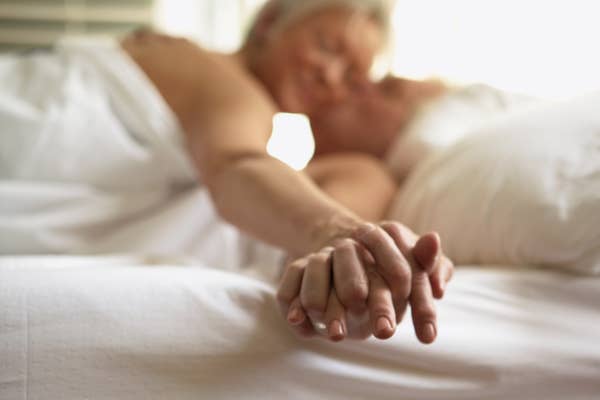 A couple holding hands in bed, partially covered by sheets, suggesting intimacy and closeness
