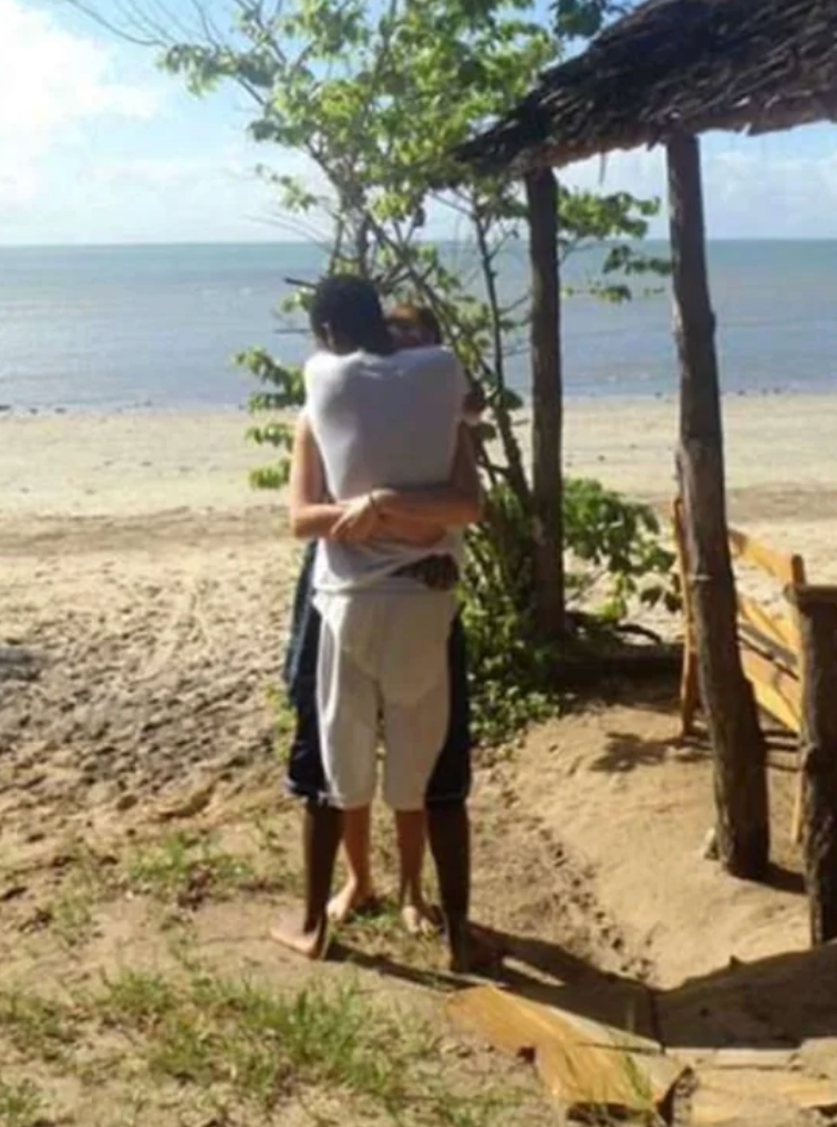 Two radical   hug connected  a sandy formation  adjacent   the ocean, nether  a rustic woody  structure  with trees nearby