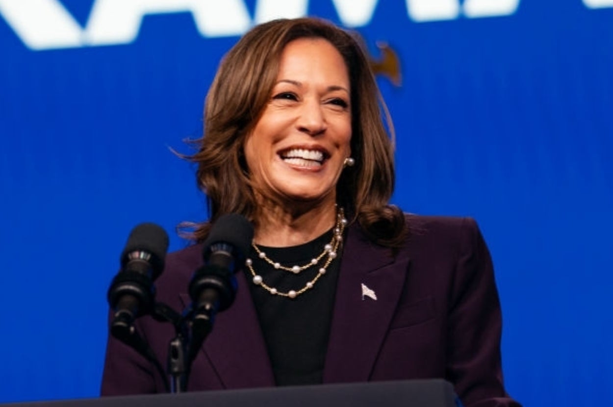 女性が演壇で話し、背後に「KAMALA」という文字が書かれている。