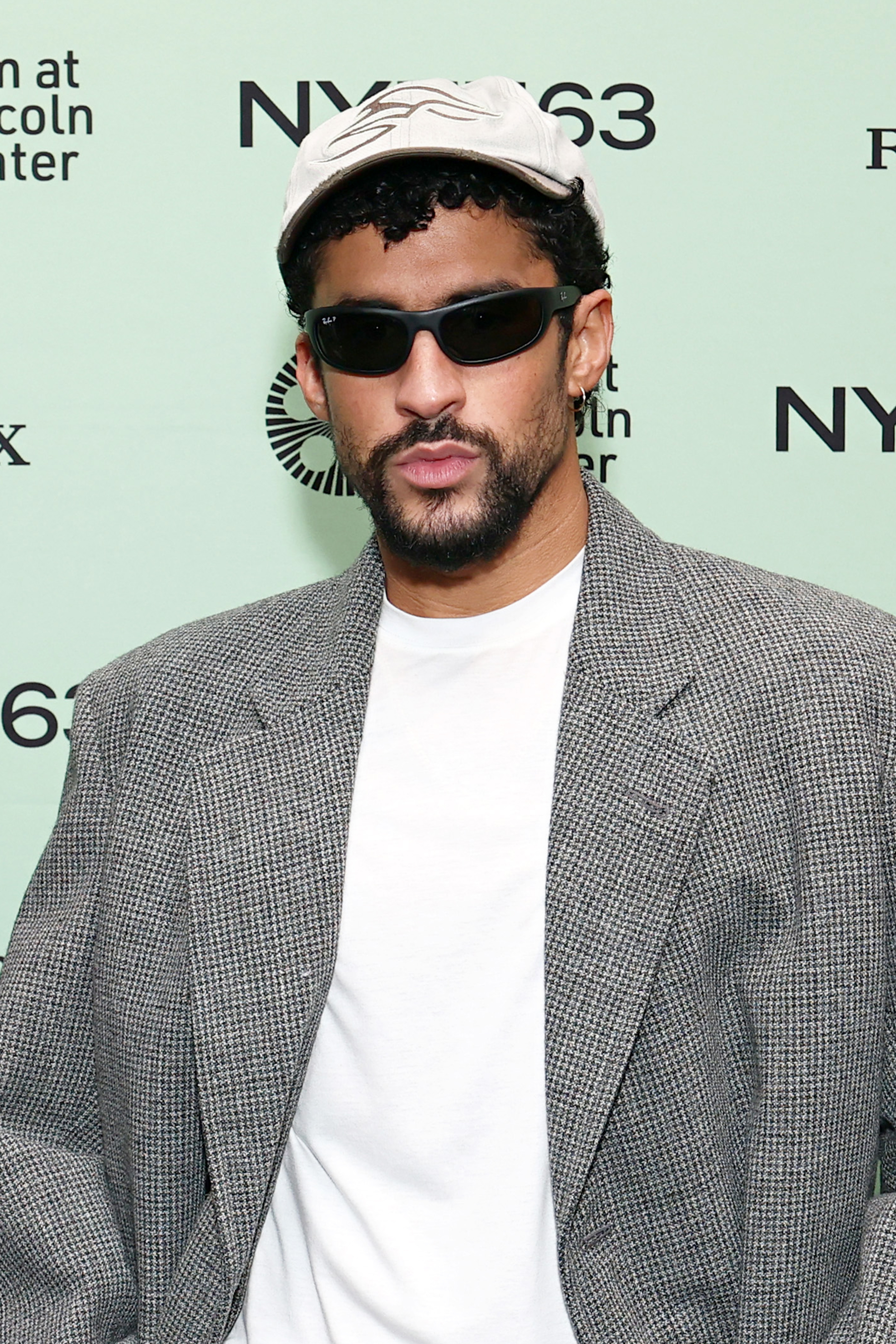 Person wearing a plaid blazer, white t-shirt, sunglasses, and cap, standing against a background with text