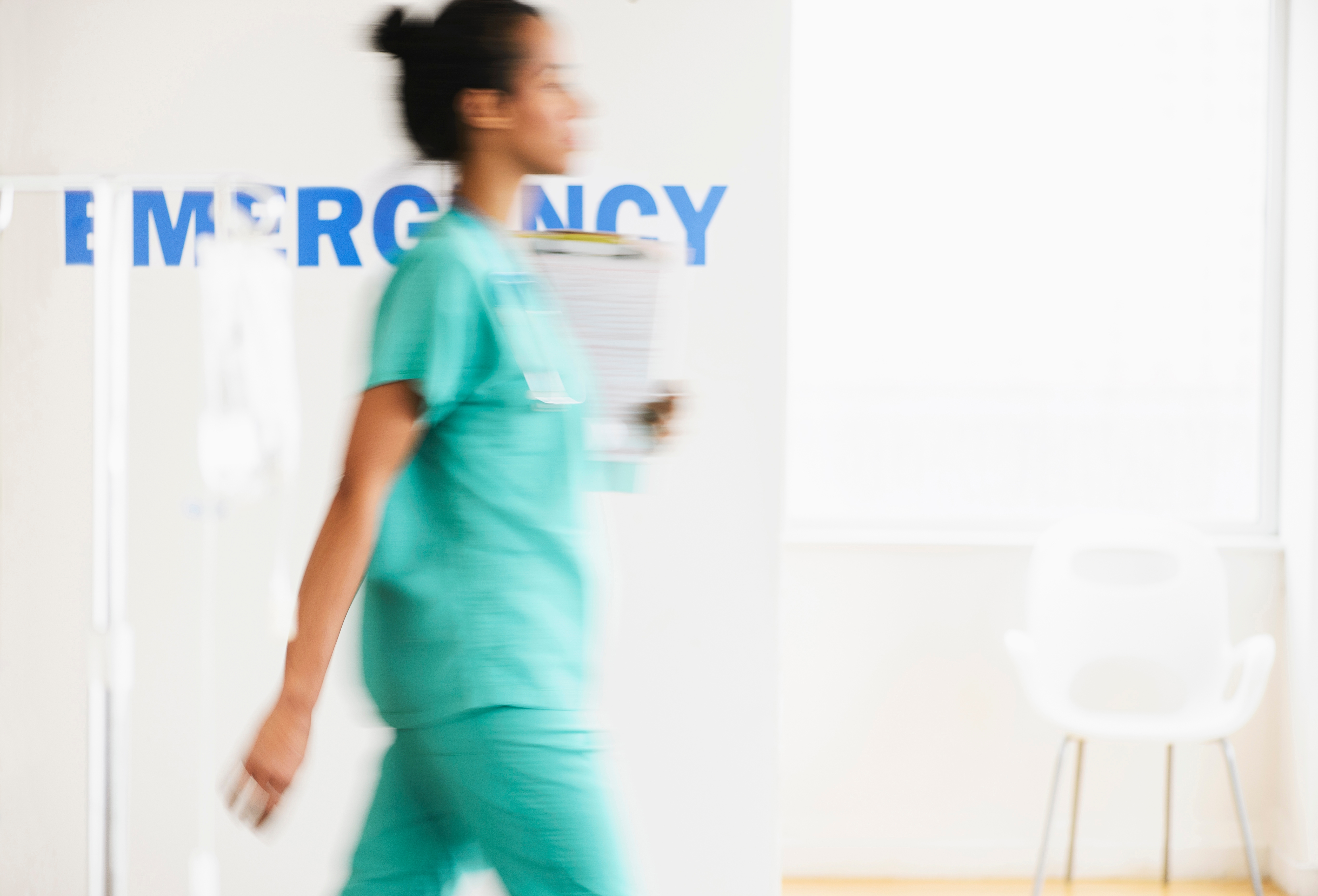 En helsepersonell i Scrubs går raskt forbi et legevaktskilt, og holder et utklippstavle