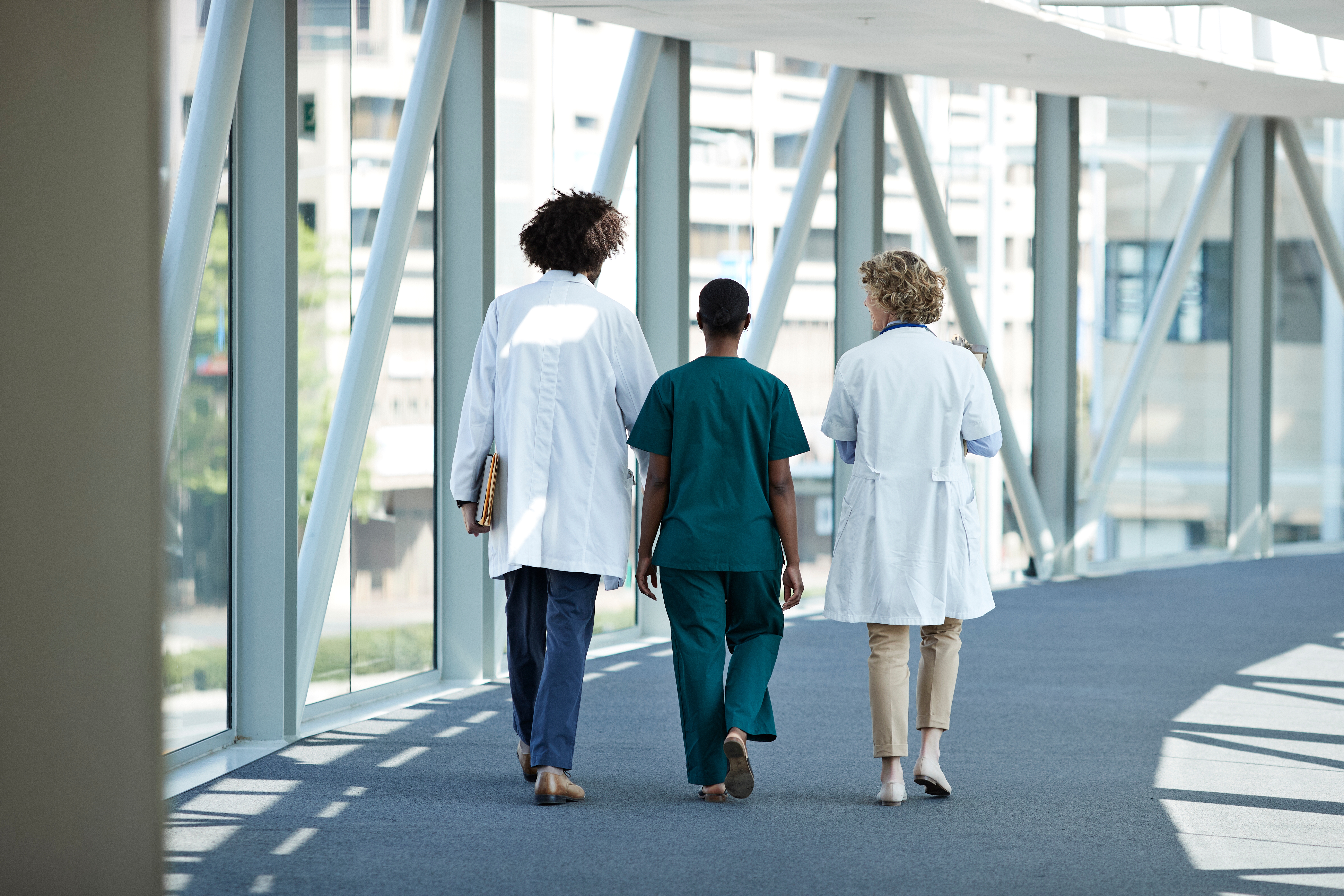 Tre medisinske fagpersoner går nedover en sykehuskorridor, to i hvite strøk og en i grønne skrubber, engasjert i samtale