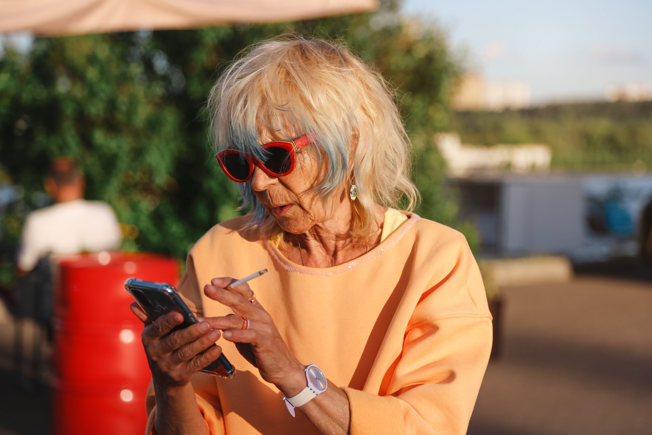Eldre kvinne iført solbriller og uformelle klær, ser på telefonen hennes mens hun holder en sigarett, utendørs
