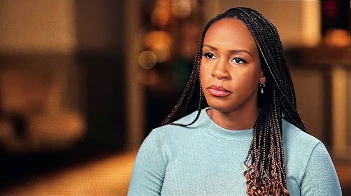 A woman with long braided hair wearing a simple, elegant blouse appears serious during an interview setting
