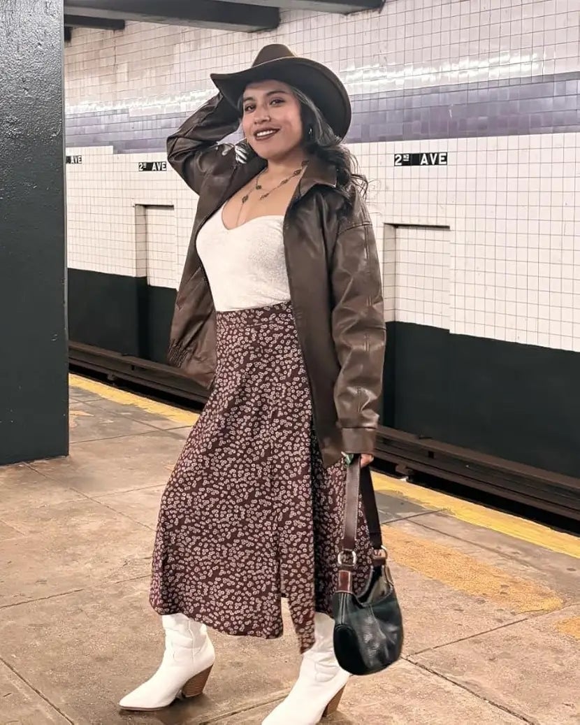 Person successful  subway presumption    wearing a brownish  jacket, achromatic  top, patterned skirt, achromatic  boots, and a cowboy hat, holding a tiny  handbag