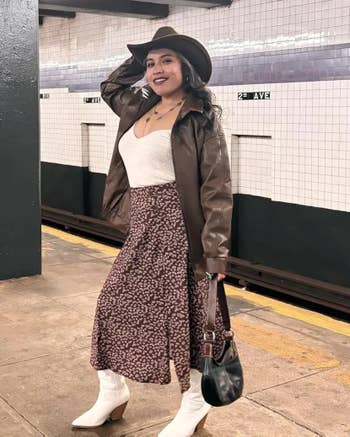 Person successful subway presumption wearing a brownish jacket, achromatic top, patterned skirt, achromatic boots, and a cowboy hat, holding a tiny handbag