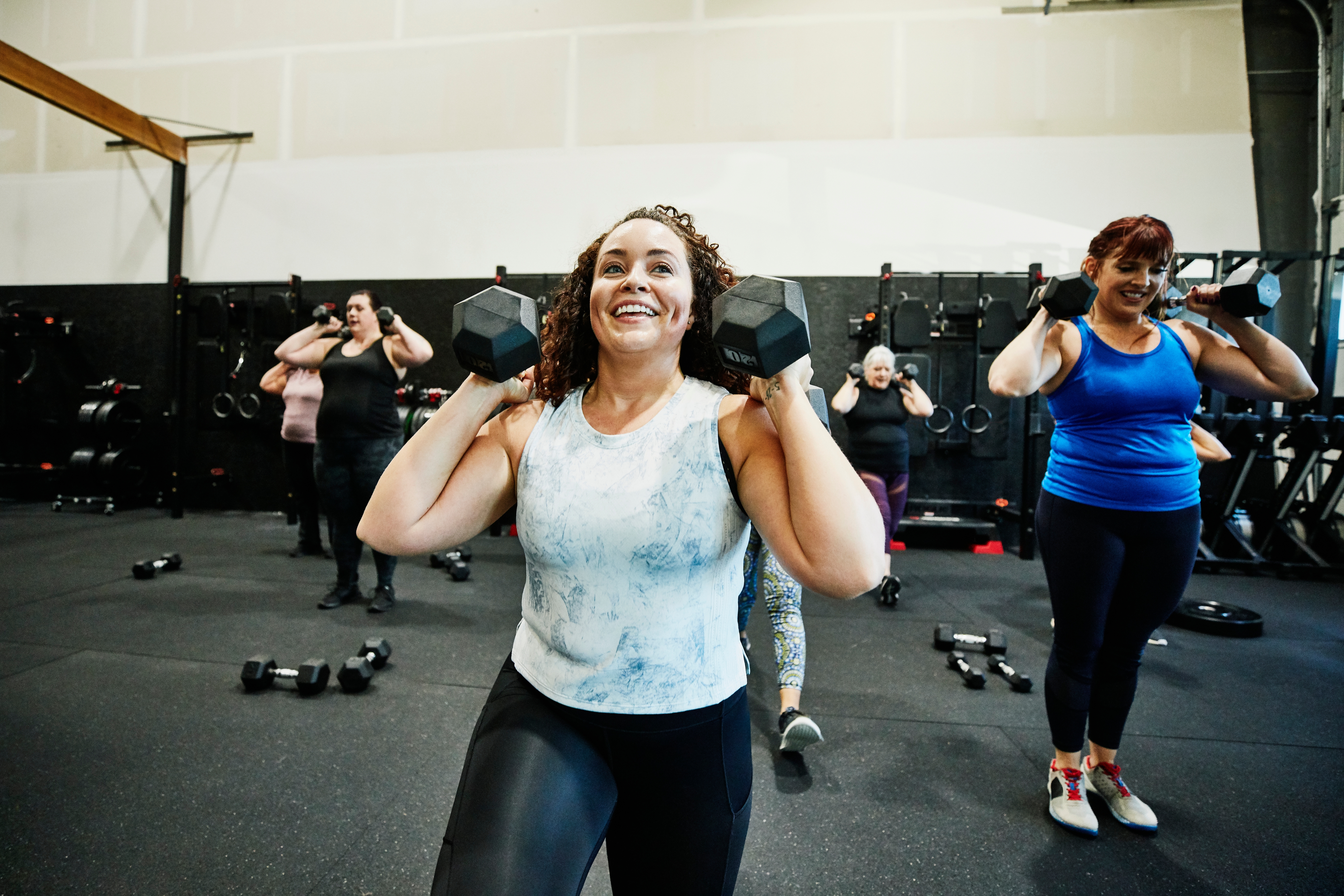 Folk i en treningstime løfter manualer, smilende og engasjerte, med ulike øvelser på gang i bakgrunnen