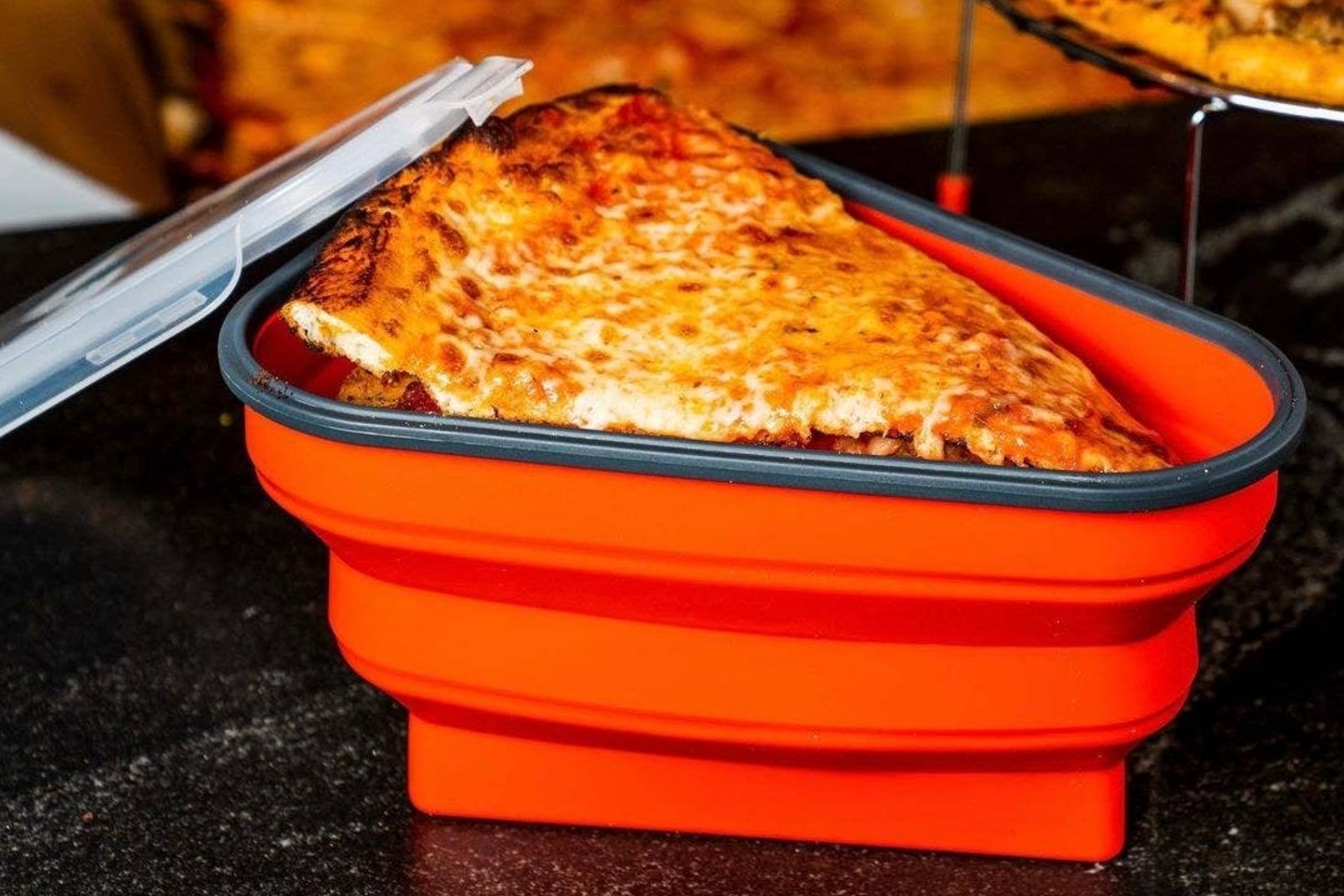 Two pizza slices stored in a collapsible orange food container with a partially open lid, displayed on a dark countertop