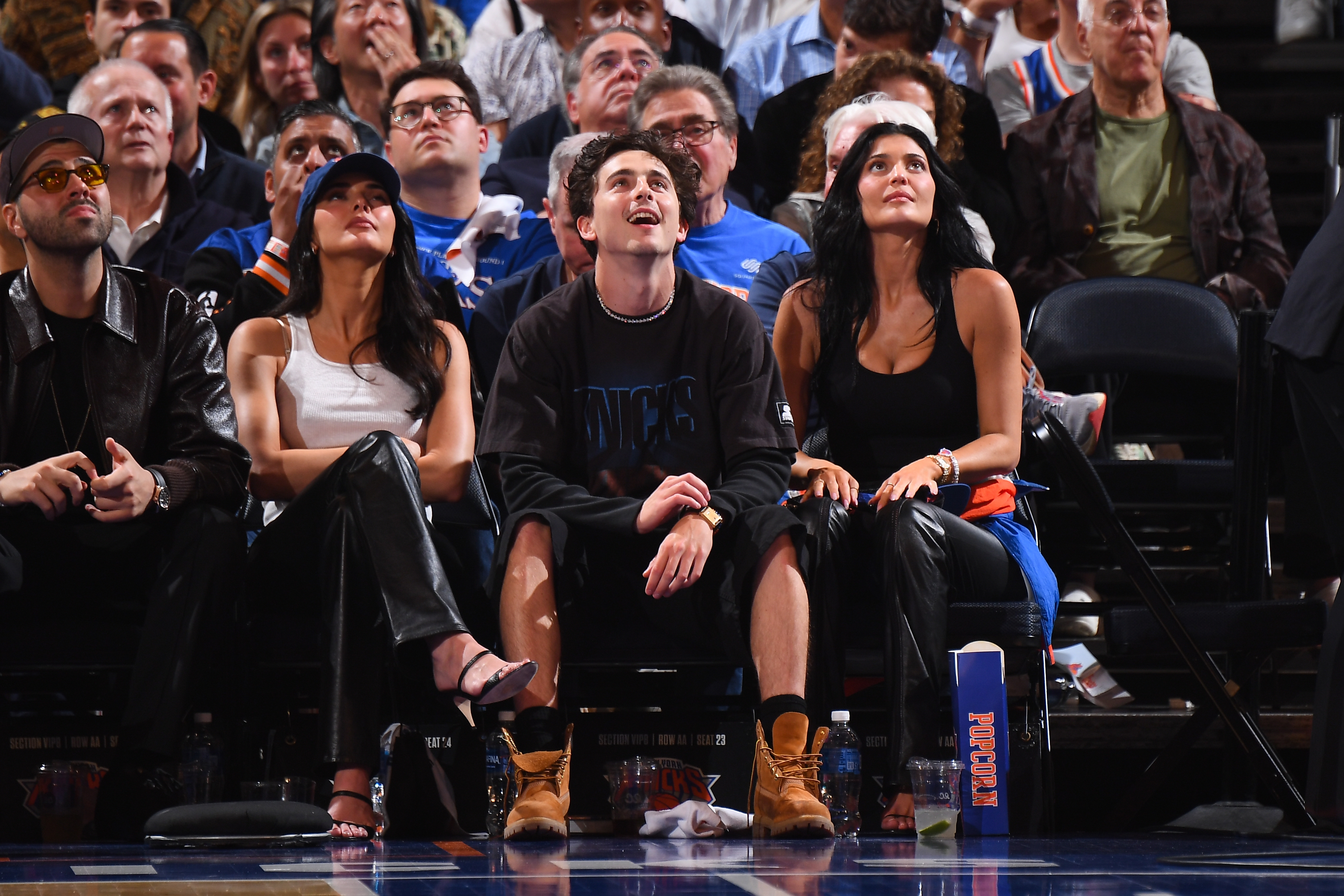Three people sit courtside at a basketball game. The two women wear stylish outfits with leather elements; the man wears boots and casual clothing