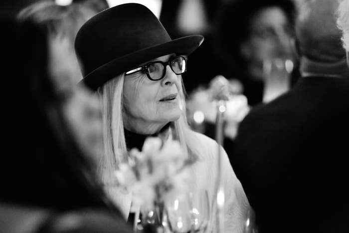 Diane Keaton in a hat and glasses at an event, surrounded by others, looking attentive